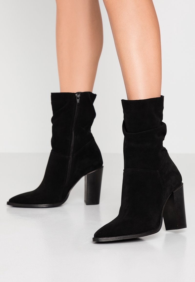 Jambes portant des bottes mi-mollet en daim noir avec des bouts pointus, des fermetures éclair latérales et des talons hauts épais sur un fond de sol blanc.