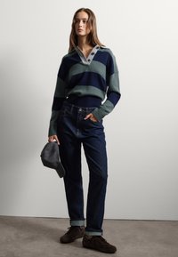 Suéter de punto a rayas en verde oscuro y marino, con un collar gris y detalles de botones. Combinado con jeans de mezclilla oscuros y una gorra gris.