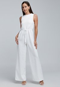 Witte jumpsuit met een smocked lijfje, wijde pijpen, een trekkoord in de taille en minimalistische bandjes. Gladde linnen textuur met een ademend gevoel.