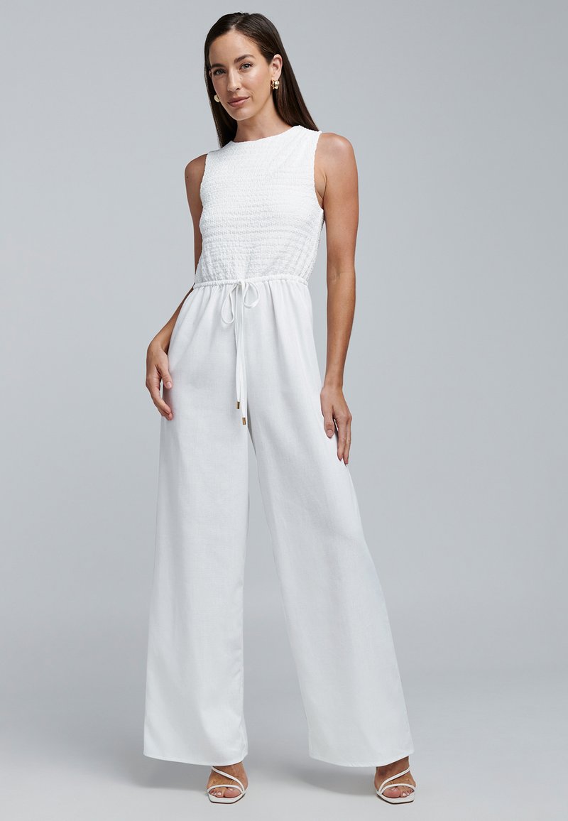 Witte jumpsuit met een smocked lijfje, wijde pijpen, een trekkoord in de taille en minimalistische bandjes. Gladde linnen textuur met een ademend gevoel.