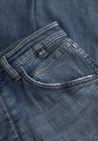 Jeans din denim albastru cu marginea buzunarului desfăcută, cusături vizibile și un nasture metalic. Textură uzată cu o finisare moale.