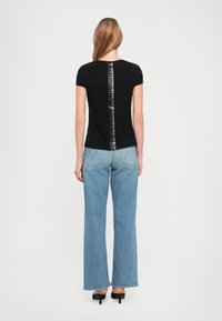 Zwarte t-shirt met korte mouwen, voorzien van een glanzend verticaal logo-detail op de rug; gecombineerd met lichtblauwe flare jeans en zwarte hakken.
