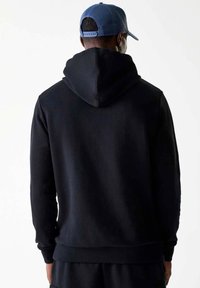 Felpa nera con cappuccio ampio, polsini e orlo aderenti. Il tessuto sembra morbido e con una texture. Indossata con un cappellino blu, vista da dietro.