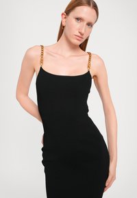 Black ribbed dress with a fitted design, featuring gold chain straps. Smooth texture with no visible patterns or embellishments.