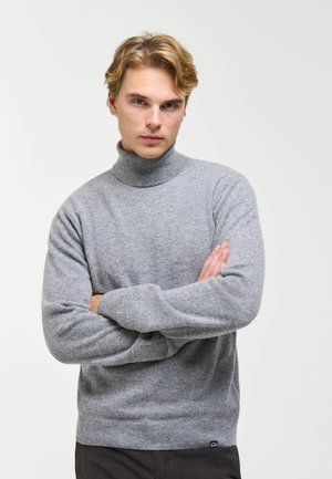 Junger Mann mit blondem Haar trägt einen grauen Rollkragenpullover, steht mit verschränkten Armen und schaut direkt geradeaus vor einem schlichten Hintergrund.