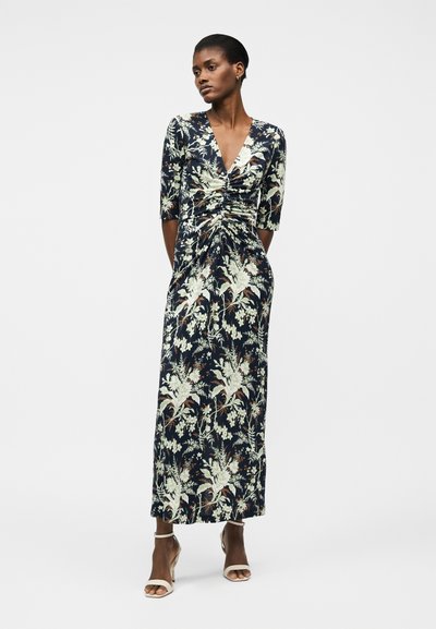 Γυναίκα που στέκεται φορώντας μια μεσάτη μπλε-ναυτικό φόρεμα με λουλουδένιο μοτίβο και 3/4 μανίκια, συνδυασμένο με λευκές ανοιχτές γόβες, μπροστά σε απλό φόντο.