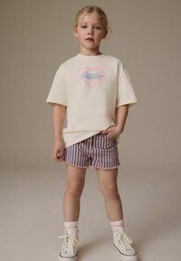 Cremfarbendes T-Shirt mit kurzen Ärmeln und einem pinken und blauen Grafikdesign, kombiniert mit gestreiften Shorts in Pink und Blau. Weiße High-Top-Sneaker runden das Outfit ab.