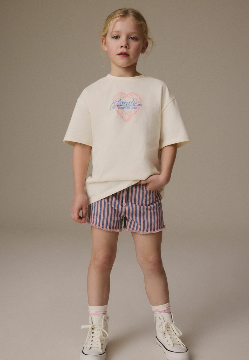 Cremfarbendes T-Shirt mit kurzen Ärmeln und einem pinken und blauen Grafikdesign, kombiniert mit gestreiften Shorts in Pink und Blau. Weiße High-Top-Sneaker runden das Outfit ab.