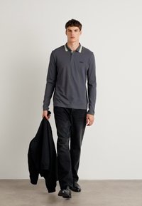 Szürke, hosszú ujjú pólóing csíkos gallérral, fekete nadrággal és cipővel párosítva. A modell egy fekete dzsekit tart. Minimalista háttér.