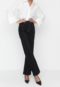 Černé džínsy s vysokým pasem a bílým prošitím, široký střih, doplněné bílou košilí na knoflíky a černými špičatými podpatky.