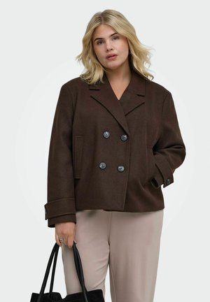 Lyshåret kvinde iført en mørkebrun dobbeltradet jakke og lyse beige bukser, der holder en sort taske med den ene hånd i jakkelommen.