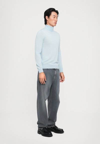 Lyseblå turtleneck genser, slim fit, myk tekstur, kombinert med løse grå jeans og svarte sko, stående mot en ensfarget bakgrunn.