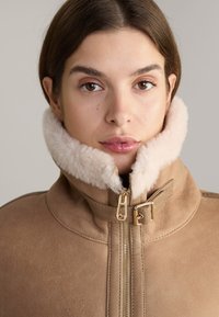 Tan suède jasje met een zachte beige schapenvacht kraag, voorzien van een gouden rits en gespdetail bij de nek, die een luxe textuur biedt.