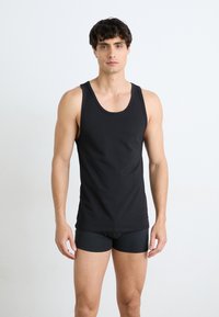 Schwarzes Tank-Top aus Baumwolle mit rundem Ausschnitt und breiten Trägern, kombiniert mit schwarzen Boxershorts, vor einem einfarbigen Hintergrund.