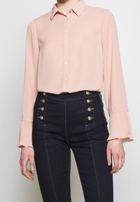 Blouse rose pâle à manches longues avec fermeture boutonnée à l'avant, présentant des poignets plissés. Pantalon taille haute bleu foncé avec des accents de boutons dorés brillants.