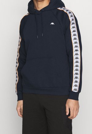 Persona che indossa una felpa con cappuccio blu navy con strisce con logo bianche e nere lungo le maniche e una tasca frontale a marsupio.