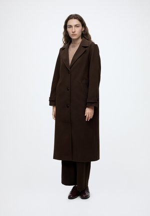 Femme aux longs cheveux bruns portant un long manteau marron foncé par-dessus un pantalon brun et des chaussures habillées, debout devant un fond blanc.