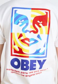 White cotton t-shirt featuring a multicolored graphic of faces, with red, blue, yellow, and turquoise shapes. "OBEY" text in bold blue.