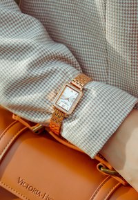 Reloj rectangular en oro rosa con cara blanca y números romanos. La correa tiene un diseño de eslabones y está adornada con pequeños cristales.