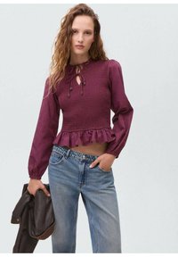 Bordeauxfarbene Bluse mit langen Ärmeln, gerüschtem Material, schlüssellochförmigem Ausschnitt und gewelltem Saum, kombiniert mit klassischen blauen Jeans und einer dunklen Jacke.