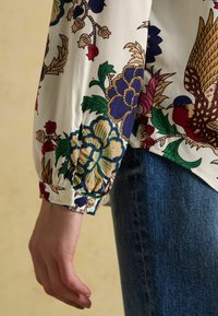 Close-up of a hand by the side wearing a floral-patterned long sleeve shirt and blue denim jeans.