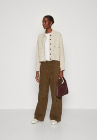 Beige linnen cropped jasje met grote bruine knopen, witte t-shirt, oversized olijfgroene cargo broek en een donker bordeauxrode handtas.