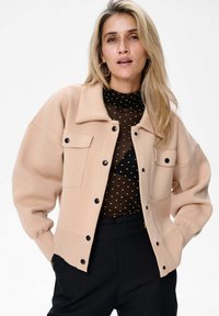 Beżowy kardigan o luźnym kroju, z dwoma przednimi kieszeniami i czarnymi guzikami. Noszony na przezroczystą bluzkę w kropki, zestawiony z ciemnymi spodniami.
