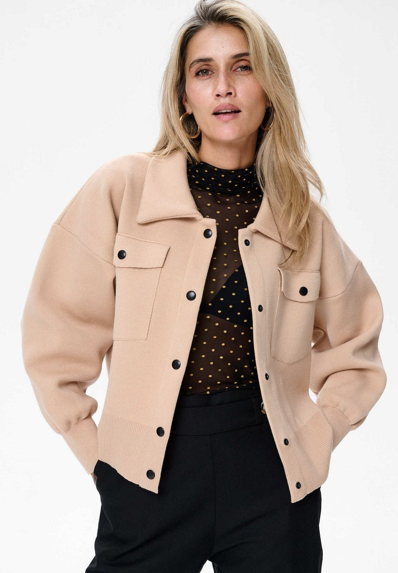 Beżowy kardigan o luźnym kroju, z dwoma przednimi kieszeniami i czarnymi guzikami. Noszony na przezroczystą bluzkę w kropki, zestawiony z ciemnymi spodniami.