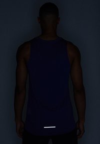 Fioletowy sportowy top na ramiączkach wykonany z lekkiego materiału, z odblaskowym akcentem z tyłu bluzki w pobliżu dolnej krawędzi i luźnym krojem.