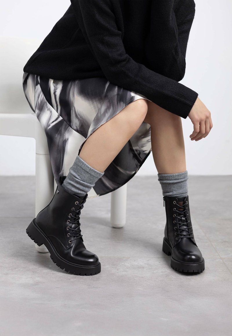 Botas de cuero negras con cordones hasta el tobillo y suelas gruesas, acompañadas de calcetines de canalé grises, junto a una falda de satén estampada sobre una superficie gris clara.