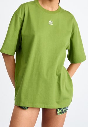 Personne portant un t-shirt vert Adidas oversize avec un petit logo blanc sur la poitrine, les mains dans le dos et un short avec un motif vert et noir.
