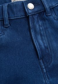 Vaqueros de mezclilla azul oscuro con una textura suave, que presentan un cierre de botón frontal y presillas para el cinturón. La vista cercana resalta los detalles de la costura.