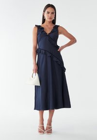 Sininen midi-mekko, jossa on röyhelökoristeet, V-pääntie, teksturoitu kangas, yhdistetty valkoiseen kolmiomaiseen laukkua ja nyöritettuihin valkoisiin sandaaleihin.