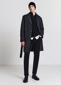 Dunkler Wollmantel mit großen Revers, Knopfverschluss, schwarzer Strickpullover, hervorblitzender weißer Kragen, schwarze Hose und glänzende schwarze Stiefel.