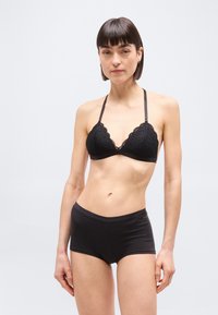 Γυναίκα με κοντά σκοτεινά μαλλιά που φοράει μαύρο δαντελένιο μπραλέτο και μαύρα σλιπ τύπου μπόισορτ, μπροστά σε απλό φωτεινό φόντο.