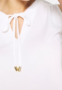 White blouse featuring a keyhole neckline with adjustable tie, accented by gold tips. Soft, smooth fabric with gentle gathers at shoulders.
