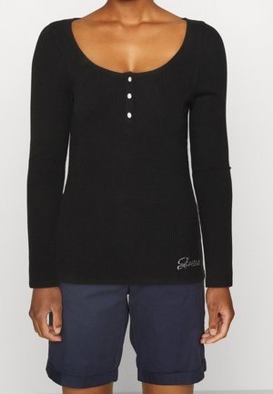 Femme portant un haut noir à manches longues côtelé avec trois boutons et un short bleu marine à revers, debout devant un fond uni.