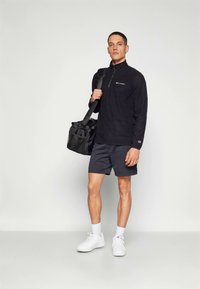 Férfi fekete hosszú ujjú pulóverben, fekete rövidnadrágban, fehér sportcipőben, vállán fekete sporttáskával, egyszínű háttér előtt.