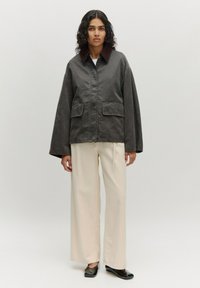 Donkergrijze jas met een bruine kraag, voorzien van een voorrits en twee grote voorzakken, gestyled met losse crèmekleurige broeken en zwarte schoenen.