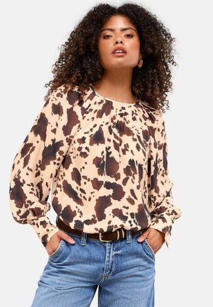 Blouse beige clair avec des taches abstraites marron foncé, dotée d'un col froncé et de manches longues, associée à un jean bleu taille haute.