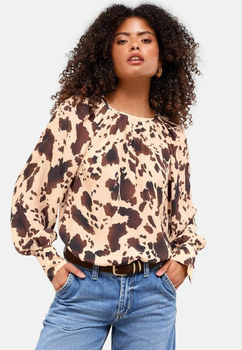 Blusa beige chiaro con macchie astratte marroni scure, con scollatura arricciata e maniche lunghe, abbinata a jeans blu a vita alta.