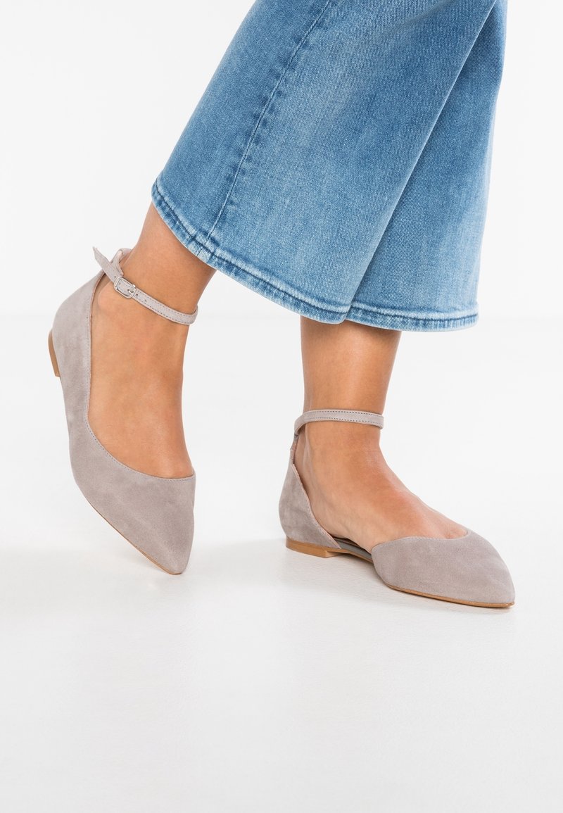 Pieds portant des ballerines pointues en daim gris avec des brides à la cheville, associées à un jean court bleu clair sur fond blanc.