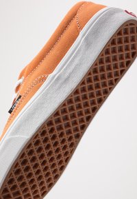 Baskets en toile orange avec lacets blancs, semelle en caoutchouc texturée et motif en nid d'abeille brun. Le côté présente des coutures blanches et une étiquette de logo.