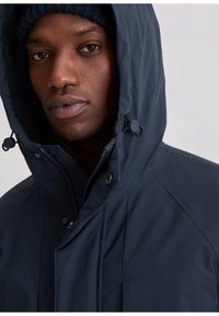 Marineblauwe capuchonjas met een hoge kraag, voorzien van een ritssluiting en drukknoopsluitingen. Gladde stof met een subtiele glans, ontworpen met getailleerde naden voor een aansluitende pasvorm.