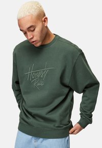 Grön sweatshirt med broderad text "Honestly Rules." Har rundad hals, långa ärmar och en avslappnad passform, tillsammans med blå jeans.