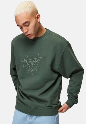 Grøn sweatshirt med broderet tekst "Honestly Rules." Har rund halsudskæring, lange ærmer og en afslappet pasform, båret med blå jeans.