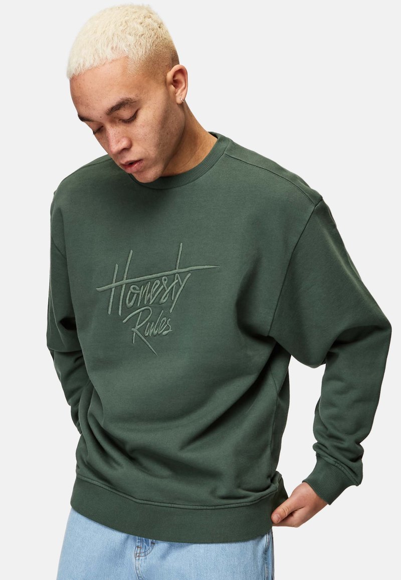 Grön sweatshirt med broderad text "Honestly Rules." Har rundad hals, långa ärmar och en avslappnad passform, tillsammans med blå jeans.