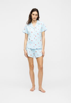 Femme debout pieds nus portant une chemise à manches courtes bleu clair boutonnée et un short assorti avec un petit imprimé floral rouge sur fond blanc.
