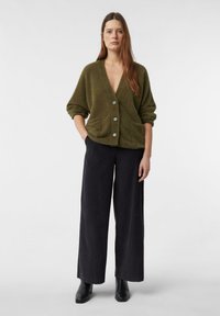 Kobieta stojąca w oliwkowozielonym, miękkim kardiganie zapinanym na guziki, szerokich czarnych spodniach i czarnych botkach na białym, jednolitym tle.