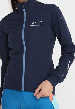 Frau trägt eine marineblaue sportliche Jacke mit hellblauem Reißverschluss und VAUDE-Logo auf Brust und Ärmel, kombiniert mit blauen Fahrradhosen.